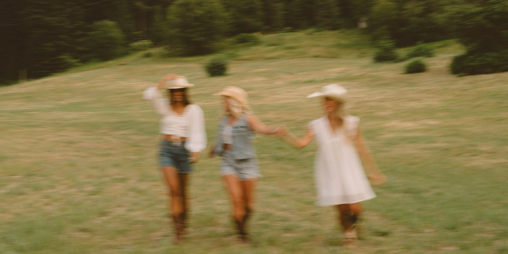Three women in summer outfits walking through a grassy field in wide-brim hats and light fabrics, representing carefree and stylish summer fashion.